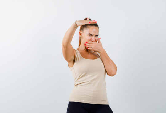 Young girl in beige top, black pants holding hand above head, covering mouth with hand and looking harried , front view.