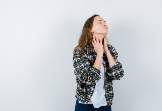 Young lady in t shirt, jacket touching skin on her neck and looking tired , front view.