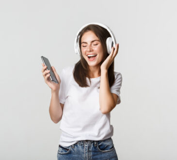 Happy beautiful brunette girl dancing and listening music in wireless headphones, holding smartphone
