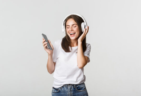 Happy beautiful brunette girl dancing and listening music in wireless headphones, holding smartphone