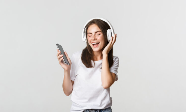 Happy beautiful brunette girl dancing and listening music in wireless headphones, holding smartphone
